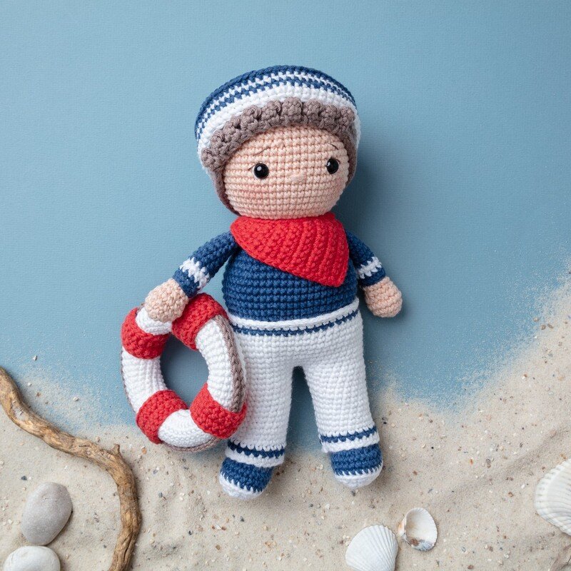 Amigurumi-Matrosenpuppe mit Mütze, rotem Halstuch und rot-weißem Rettungsring auf blauem Hintergrund mit Sand und Muscheln