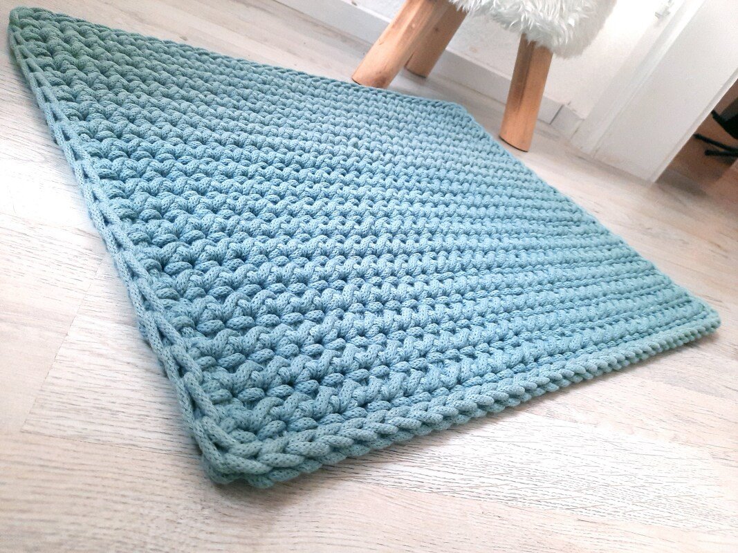 Hellblaue, rechteckige Häkelmatte mit Reliefmuster auf hellem Holzboden.