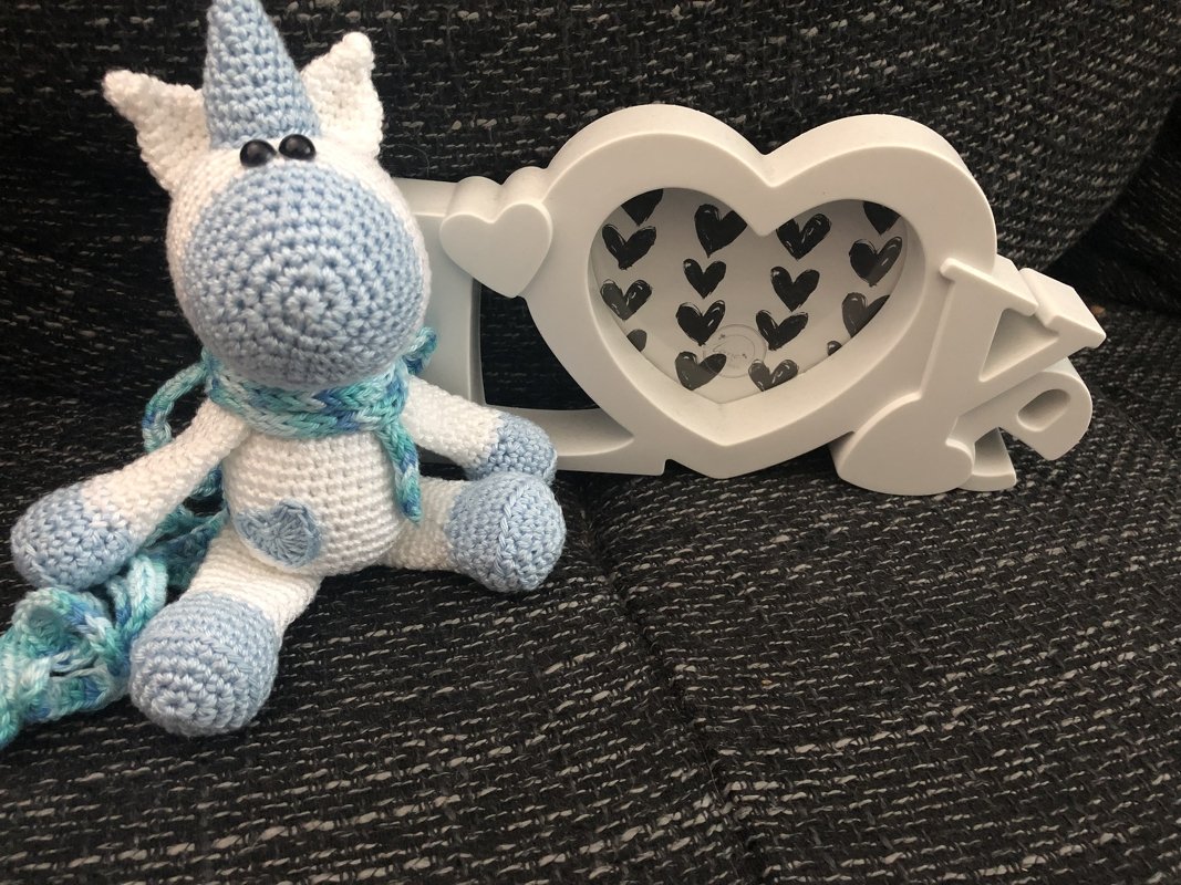 Weißes und hellblaues gehäkeltes Einhorn-Amigurumi mit blau-türkisem Schal neben einem weißen Herzrahmen.
