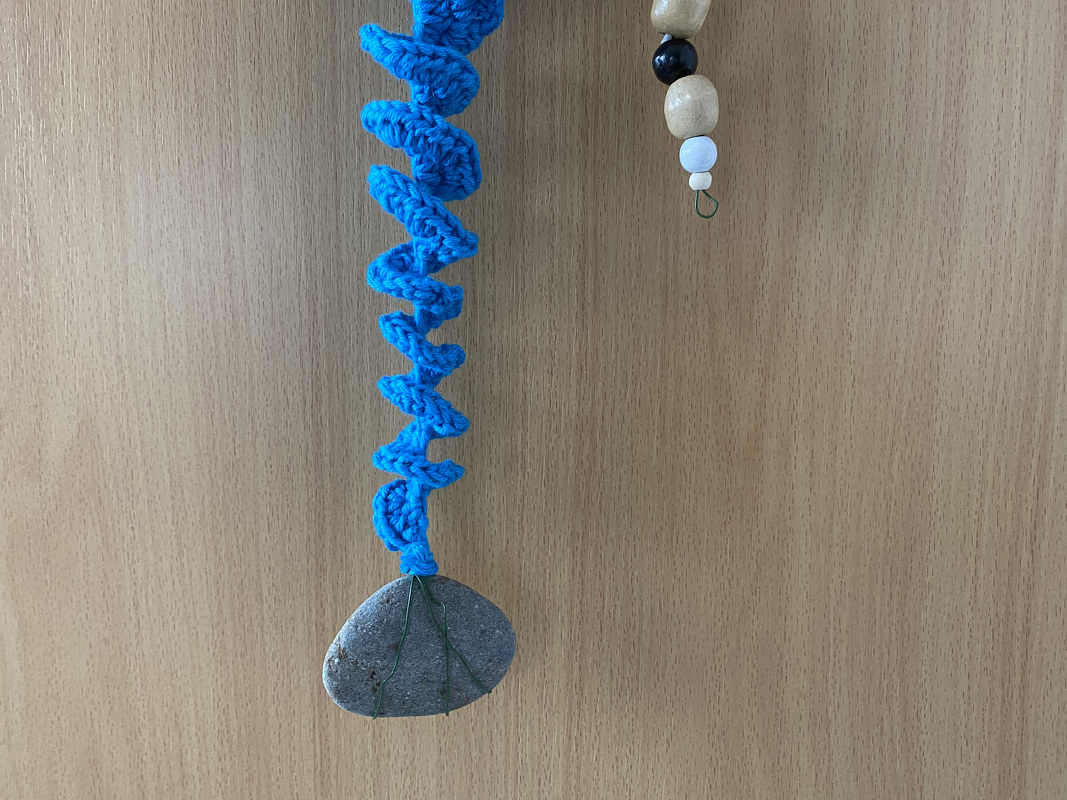 Blaue gehäkelte Spirale mit grauem Steinanhänger vor hellbrauner Holzwand, rechts Holzperlen erkennbar.