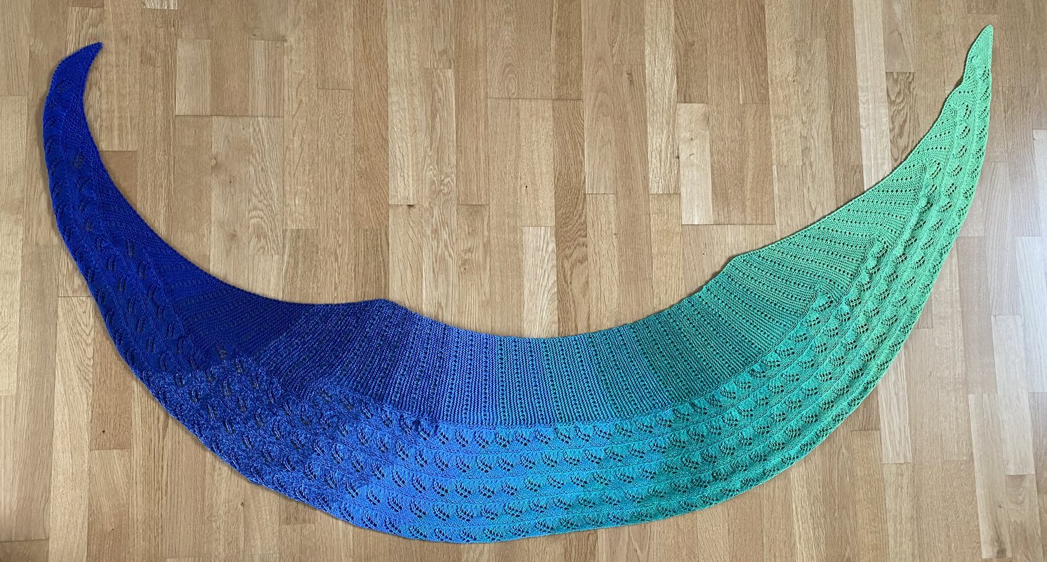 Sichelförmiger gestrickter Schal mit Farbverlauf von Blau zu Türkis und feinem Lochmuster auf hellem Holzboden