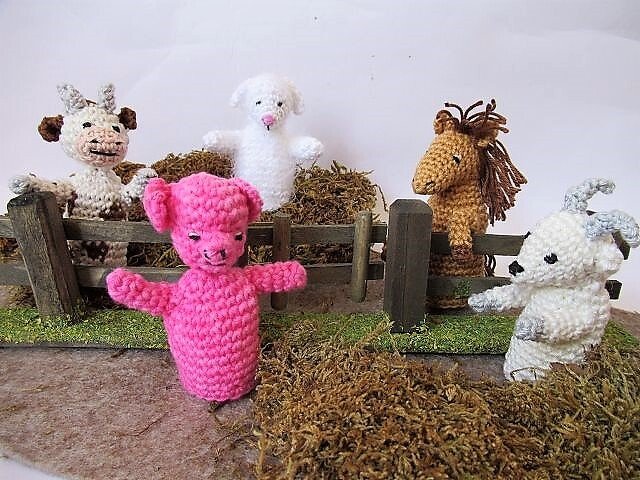 Fünf gehäkelte Amigurumi-Fingerpuppen – rosa Schwein vorn, Kuh, Schaf, Pferd und Ziege vor einem Holzzaun
