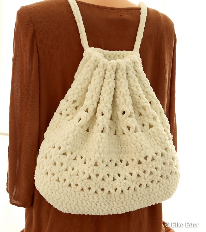 Back view of a cream chunky crochet backpack with braided straps and drawstring closure worn over a brown top