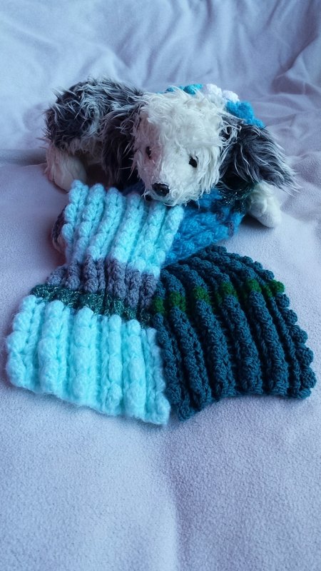 Gehäkelter Kinderschal mit horizontalen Streifen in Hellblau, Dunkelblau, Grau und Grün auf einer hellen Decke neben einem Plüschtier