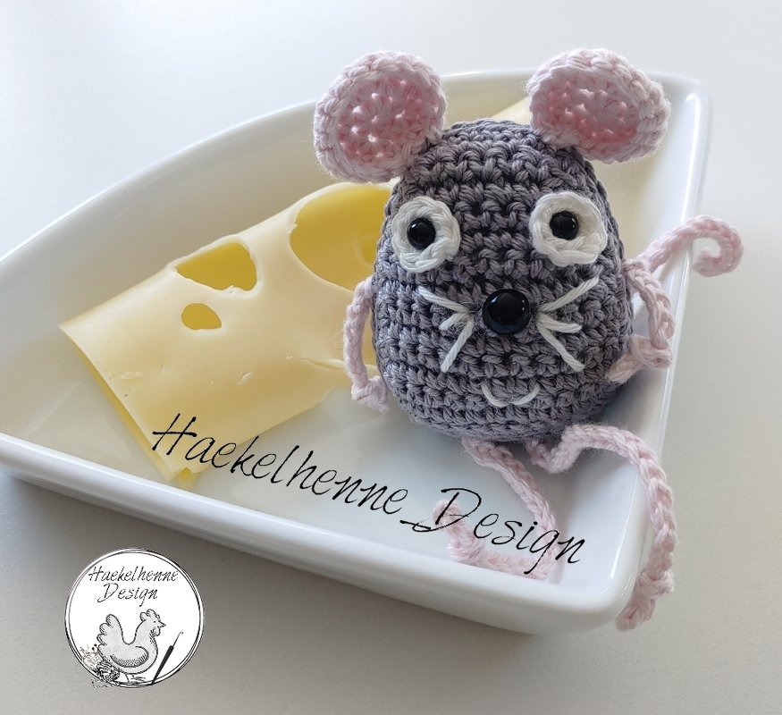 Gehäkelte graue Amigurumi-Maus mit rosa Ohren und Beinen neben einer Käsescheibe auf einer weißen Schale.