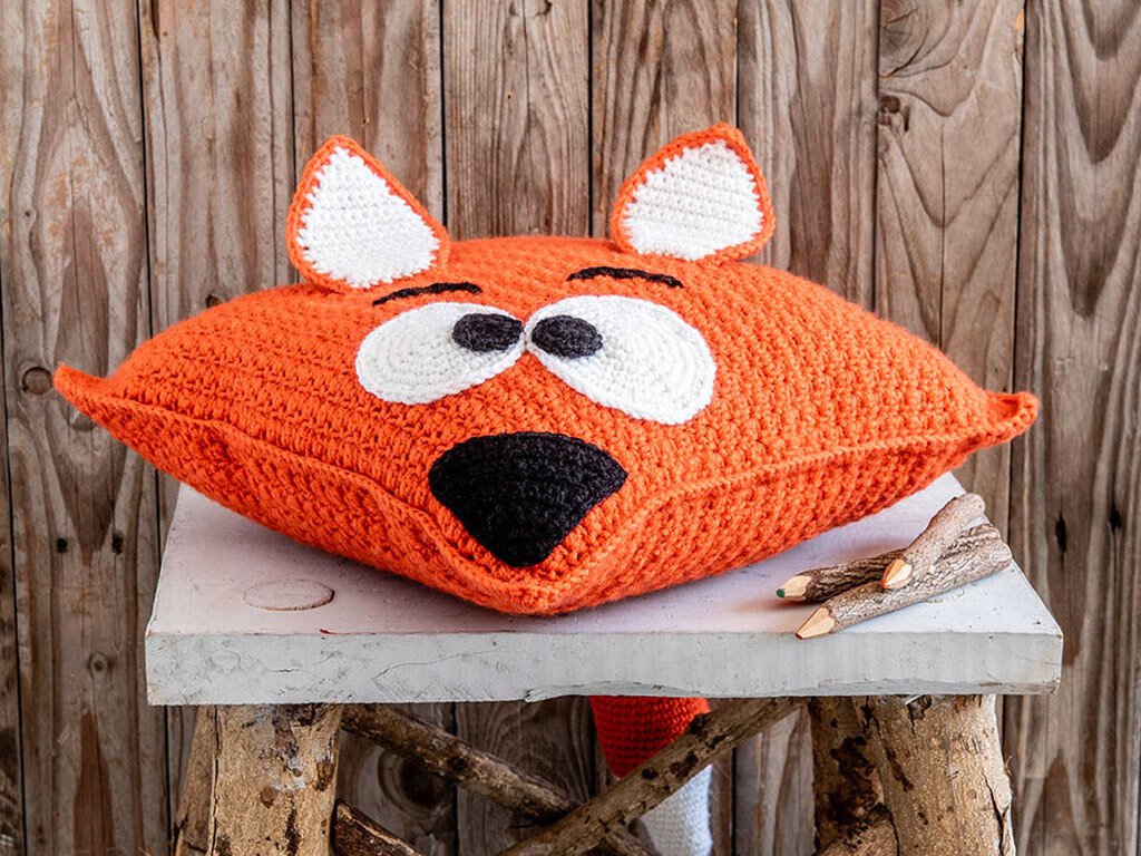 Gehäkeltes orange Fuchskissen mit weißen Augen, schwarzer Nase und Ohren auf Holzuntergrund