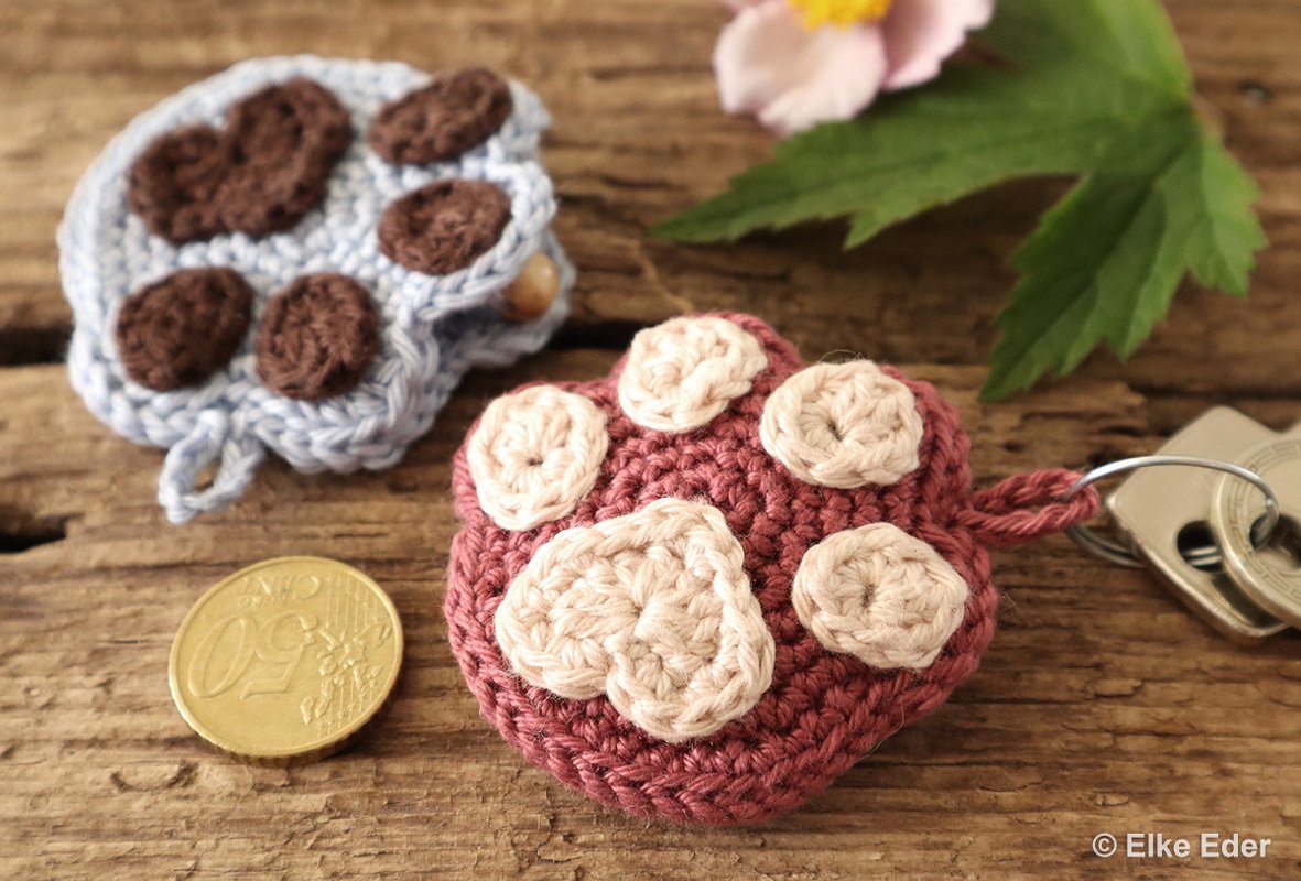 Round crocheted paw keychain in dusty rose with cream pads, a coin and keys nearby on a wooden table, with a blue paw in background