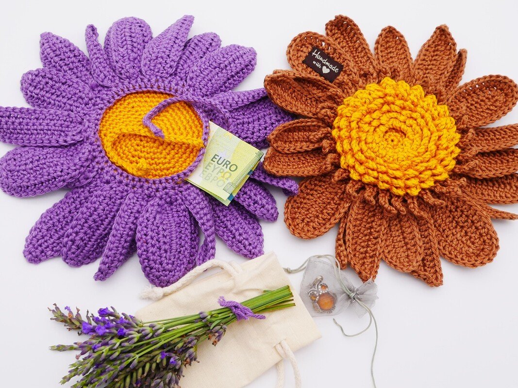 Zwei gehäkelte Blumen (lila und braun), gefalteter Geldschein, Lavendel und Schmuckbeutel auf weißem Grund.