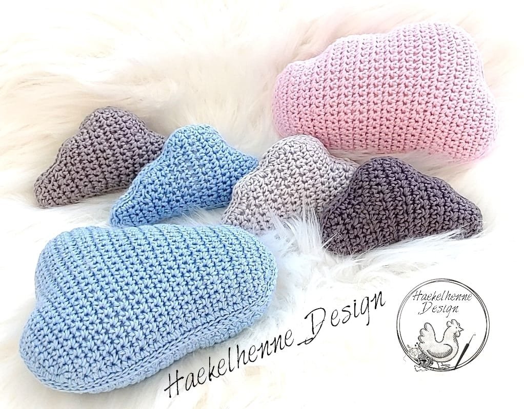 Gehäkelte Wolkenkissen in Hellblau, Rosa und Grau auf weißem, flauschigem Untergrund.