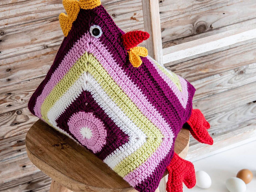 Quadratisches gehäkeltes Hühner-Kissen in Violett, Rosa, Hellgrün und Weiß mit gelbem Kamm und rotem Schnabel auf Holzstuhl