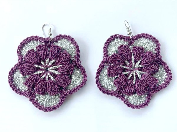Paar gehäkelter Blumenohrringe in Lila und silberfarbenem Garn mit Ohrhaken auf hellem Hintergrund.