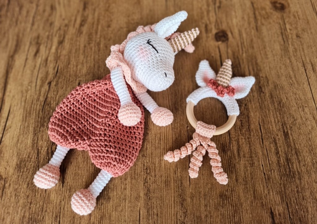 Gehäkeltes Einhorn-Schmusetuch in Weiß und Altrosa neben einem passenden Beißring mit Holzring auf Holzoberfläche