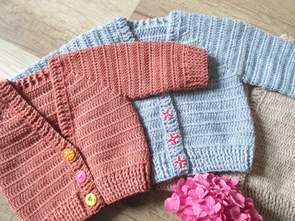 Drei gehäkelte Babyjacken in Hellblau, Terrakotta und Beige auf Holz, dekoriert mit rosa Blüten.