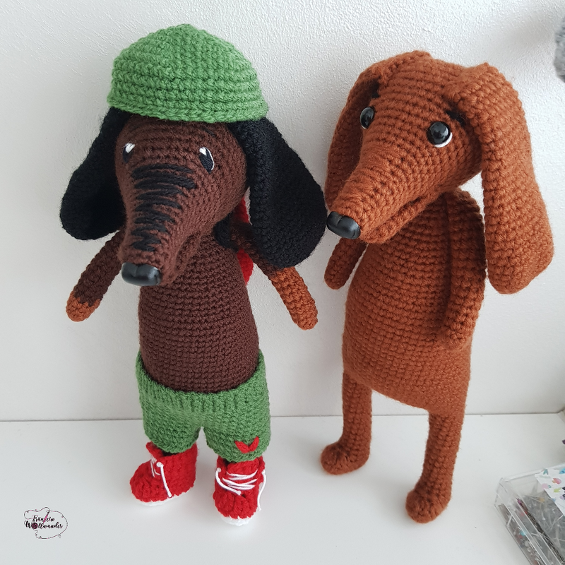 Zwei gehäkelte Dackel-Amigurumi in Braun, links mit grüner Mütze und roten Schuhen, rechts schlicht braun