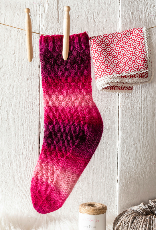 Gestrickte Damensocke in Magenta- und Pinktönen mit dekorativem Strukturmuster, aufgehängt mit Holzklammern an einer Wäscheleine.