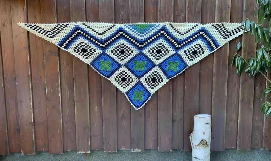 Gehäkeltes dreieckiges Tuch aus Granny‑Squares und Zickzackstreifen in Blau, Weiß, Grün und Schwarz an einer Holzlattenwand
