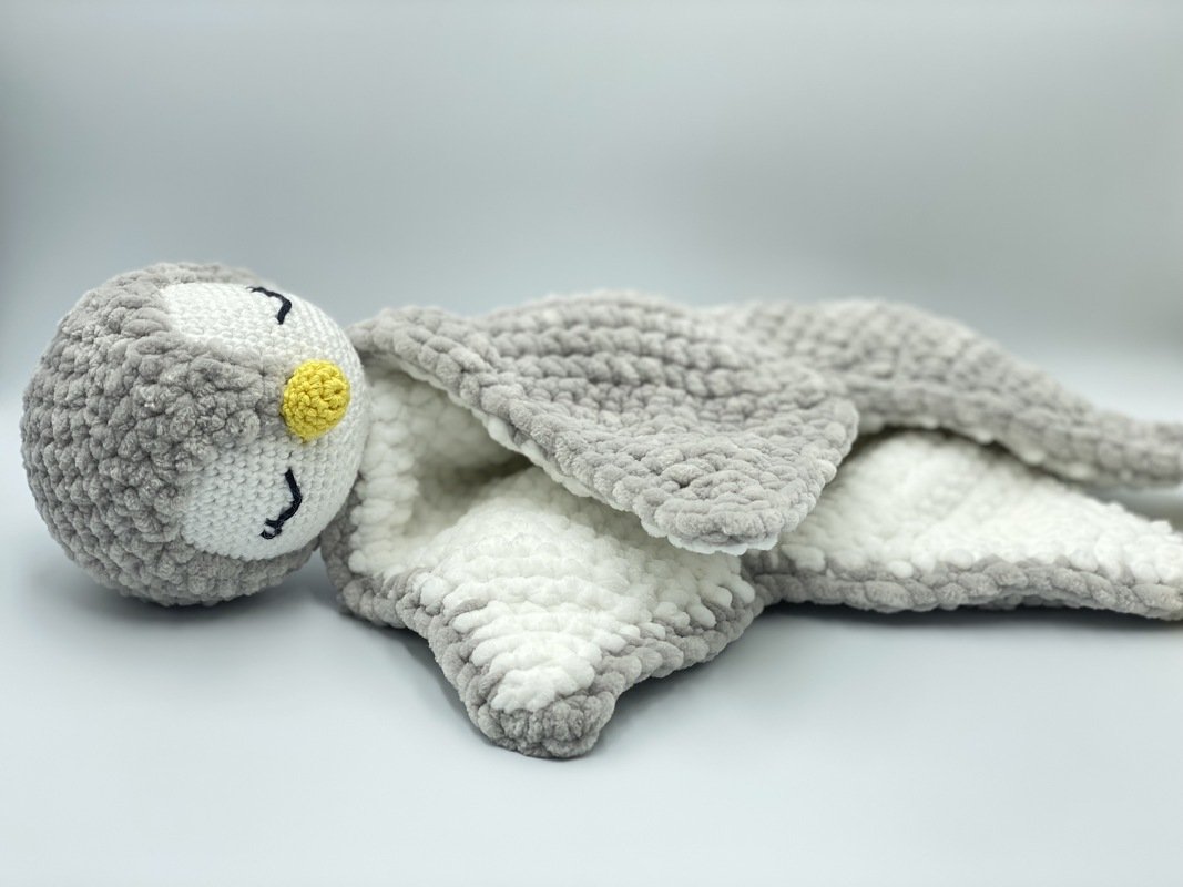 Gray and white crocheted baby snuggle blanket with a plush round head, embroidered closed eyes and yellow nose
