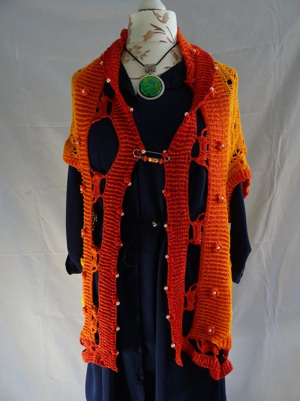 Schneiderpuppe trägt orange‑rote, gehäkelte Weste mit offenem Lochmuster und Perlen über einem dunklen Kleid.