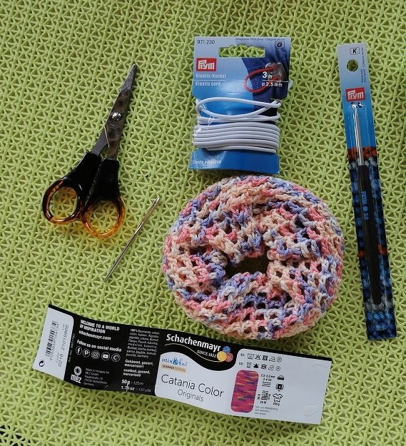 Gehäkelter Scrunchie, Garnetikett, Häkelnadel in Verpackung, Sticknadel, Schere und elastisches Band auf grünem Untergrund