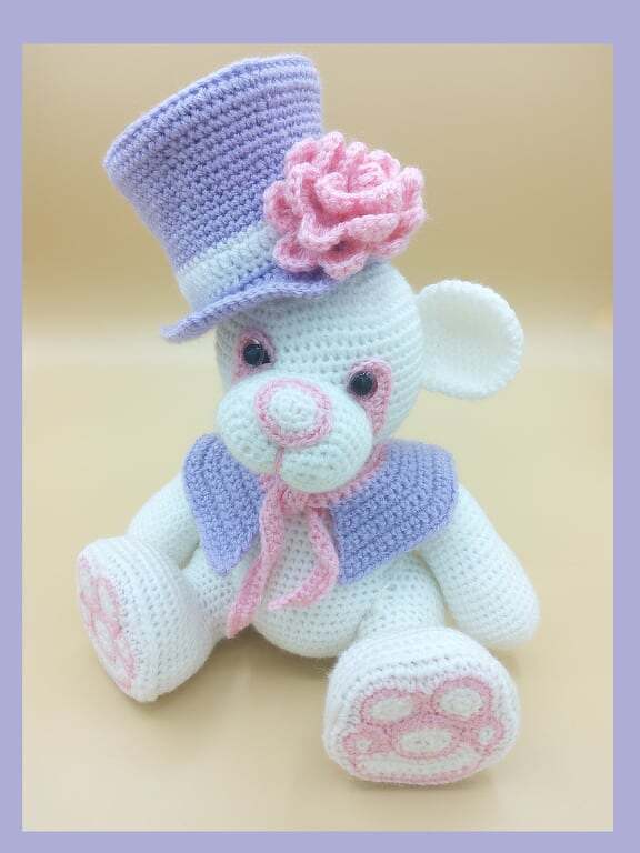 Weißer Amigurumi-Teddybär mit lila Zylinderhut und rosa Rose, sitzend