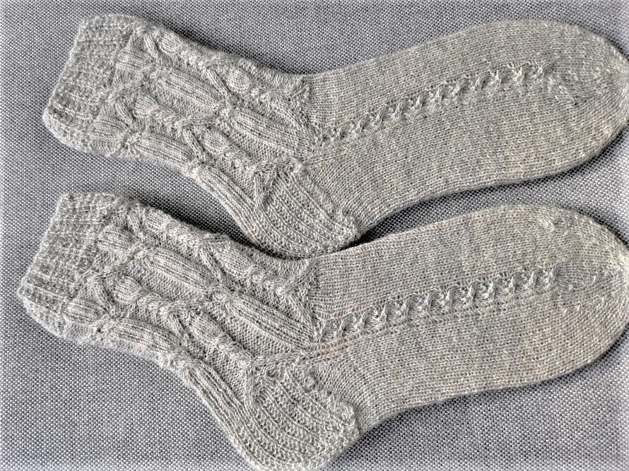 Zwei graue, handgestrickte Damensocken mit Zopf- und Lochmuster, nebeneinander auf hellgrauem Stoff.