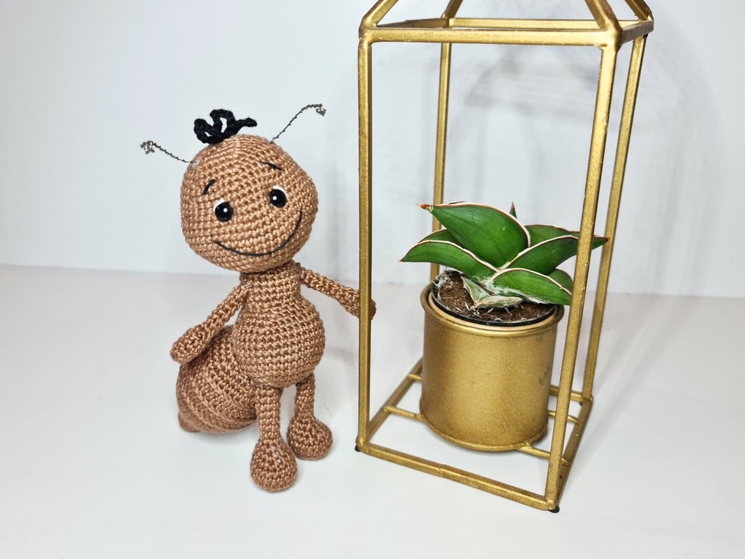 Gehäkelte braune Amigurumi-Ameise mit lächelndem Gesicht und Drahtfühlern neben goldener Pflanzen-Deko