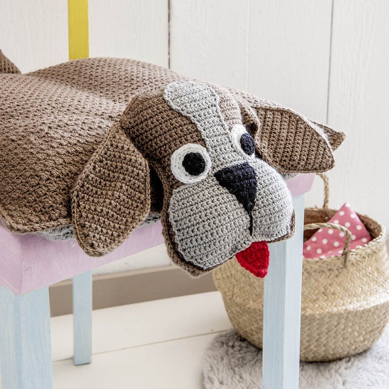 Gehäkeltes Hundekissen in Braun und Grau mit roter Zunge auf einer Bank.
