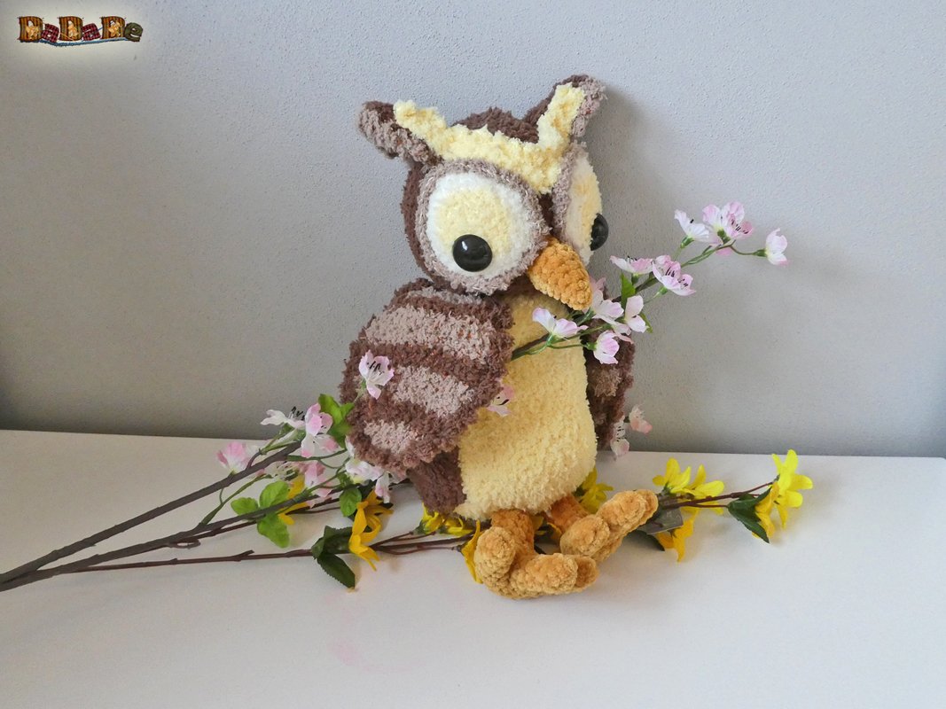 Gehäkelte Eule in Braun, Beige und Gelb mit schwarzen Augen und orangefarbenem Schnabel, umgeben von künstlichen Blüten