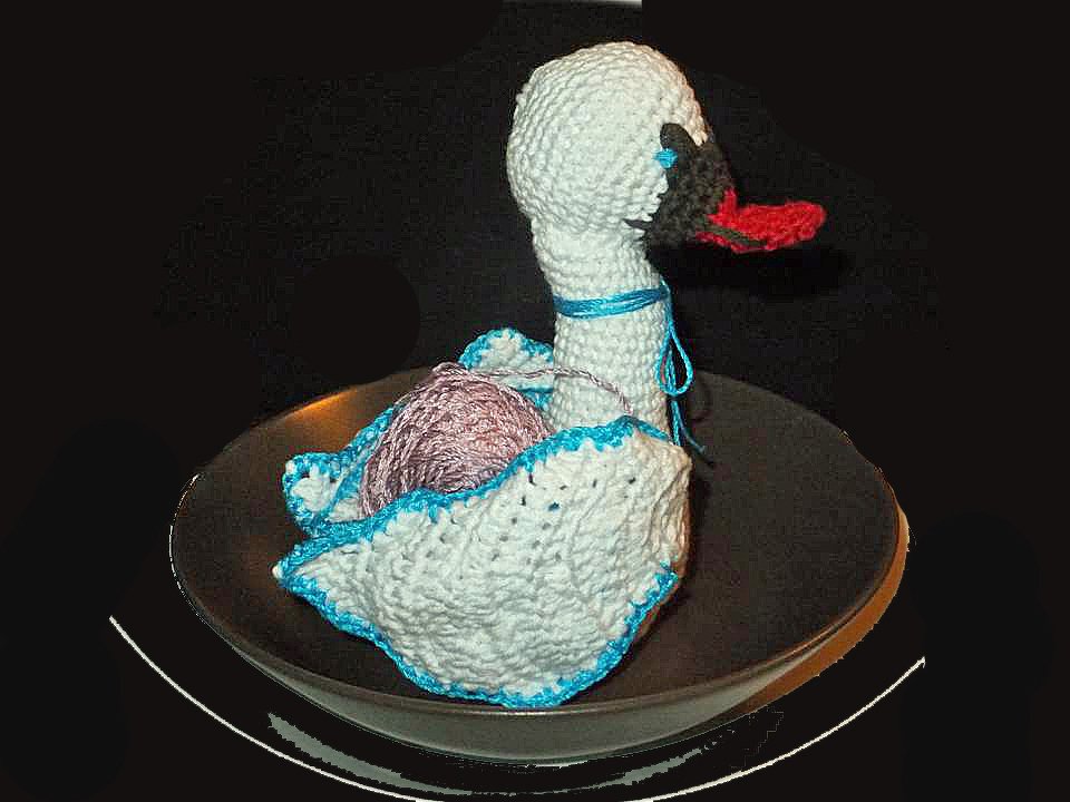 Weiß gehäkelter Schwan mit rotem Schnabel und blauer Zierkante, der ein rosafarbenes Garnknäuel im Körper hält.