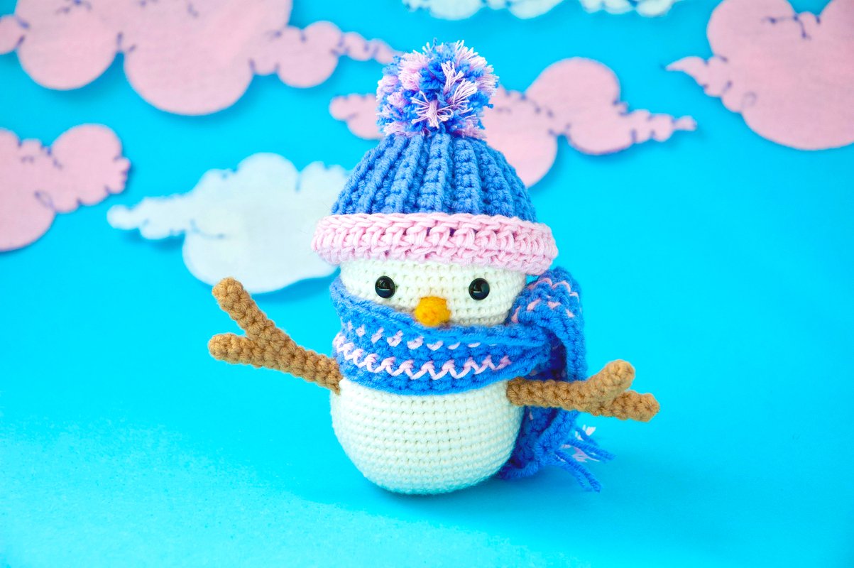 Crochet amigurumi snowman with blue pom-pom hat and matching scarf on a pastel cloud background