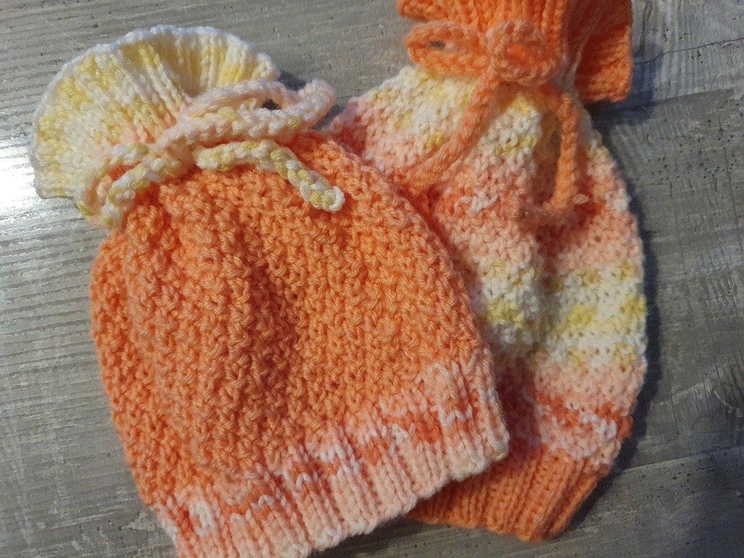 Nahaufnahme zweier gestrickter Babymützen in orange-pfirsich-Tönen mit Rippenbündchen, Strukturmasche und Bindekordel