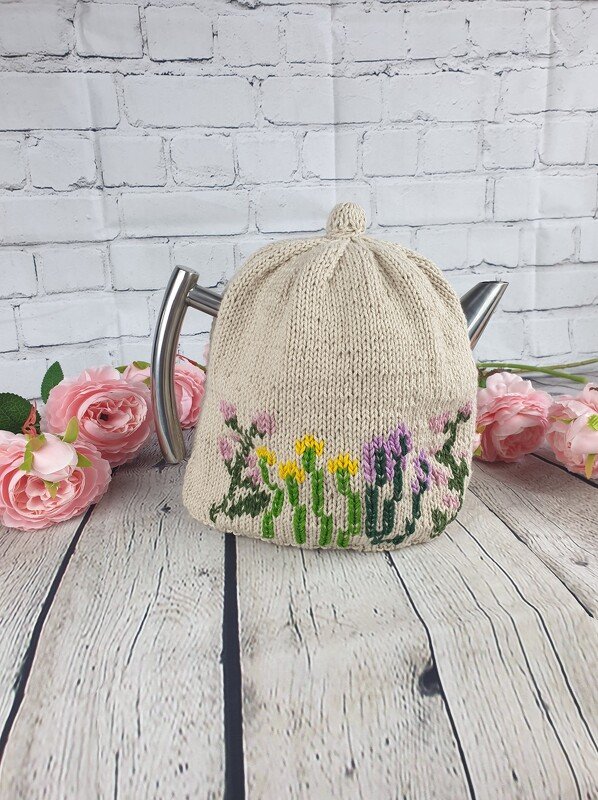 Beigefarbener gestrickter Teewärmer mit gelben, lila und grünen Blumen auf Teekanne, vor weißer Ziegelwand und rosa Rosen.