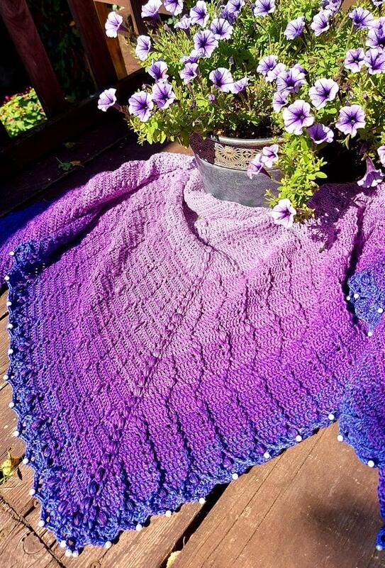 Purple ombré crochet triangle shawl with lacy stitch pattern and beaded scalloped edge laid flat beside a pot of petunias on a wooden deck