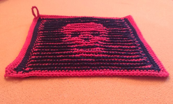 Quadratischer gestrickter Topflappen in Dunkelblau mit pinkem Totenkopf, Umrandung und Aufhängeschlaufe.