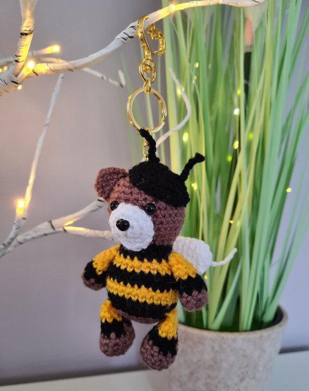 Gehäkelter Amigurumi-Teddy im Bienenkostüm als Schlüsselanhänger mit goldfarbenem Ring, hängend vor einer Topfpflanze