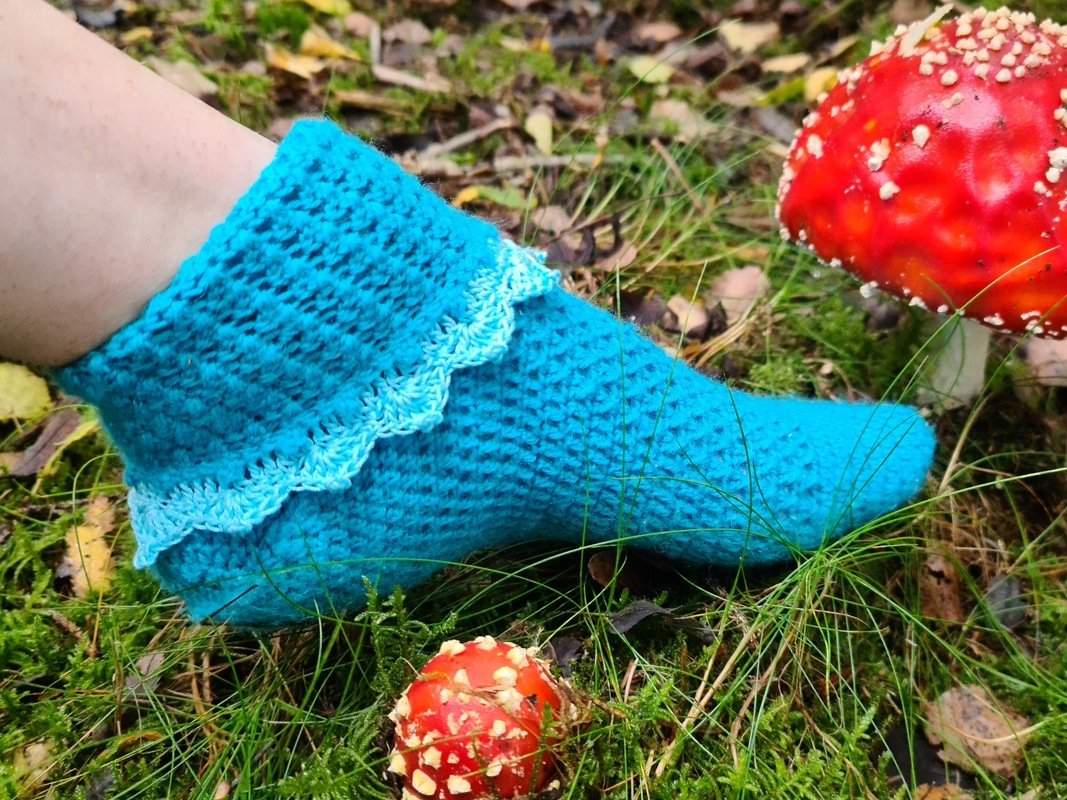 Türkise gehäkelte Socke mit hellblauer Wellenkante auf moosigem Waldboden neben roten Fliegenpilzen.
