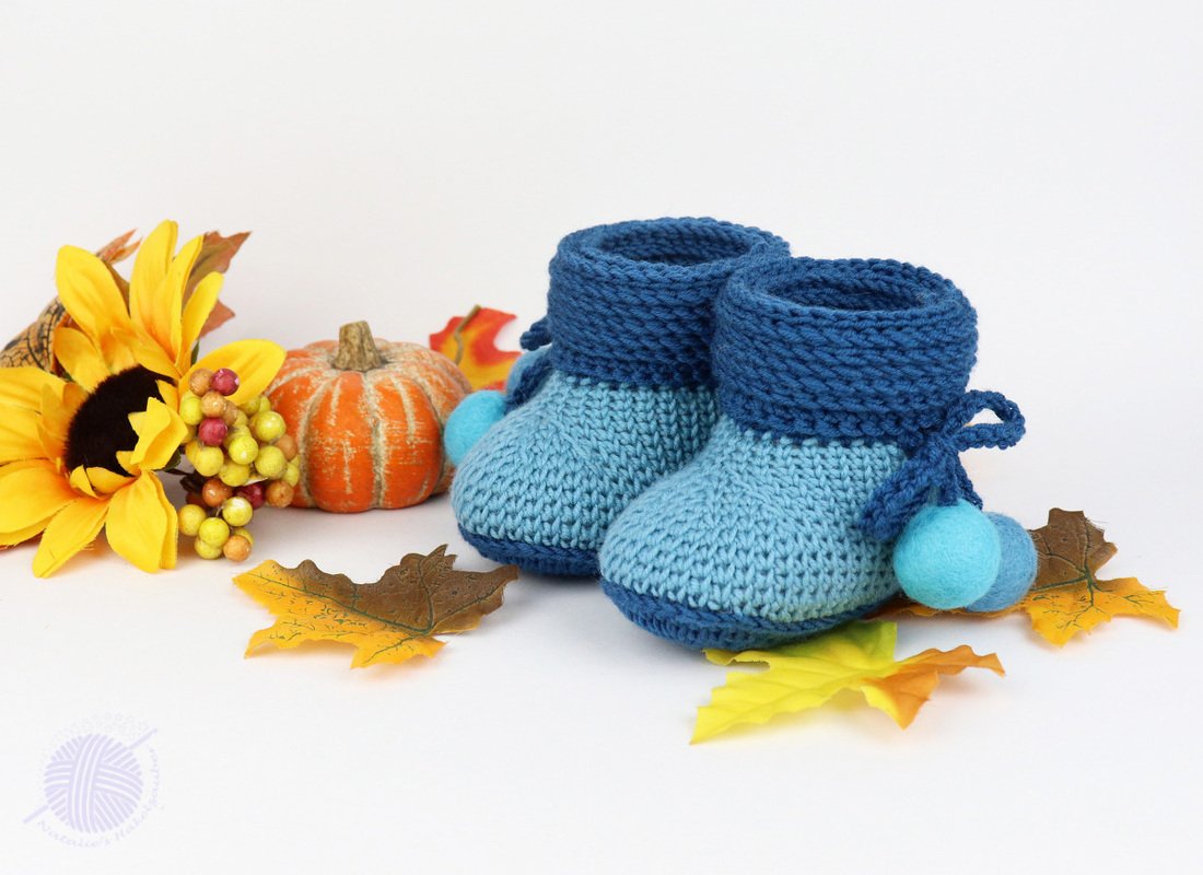 Soft blue crochet baby booties with felt pom-poms beside a pumpkin, sunflower, and fall leaves