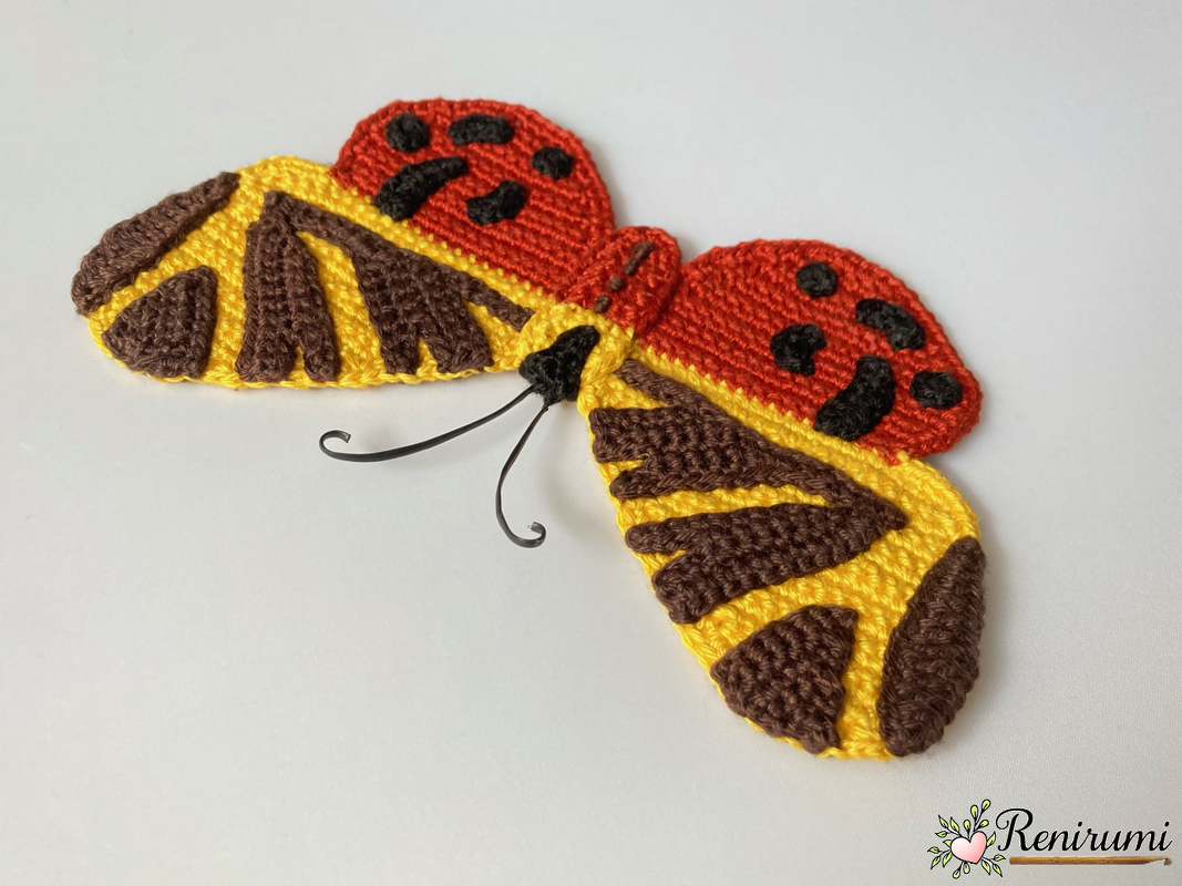 Crochet butterfly applique with red, yellow and brown wings and thin wire antennae on a white background.
