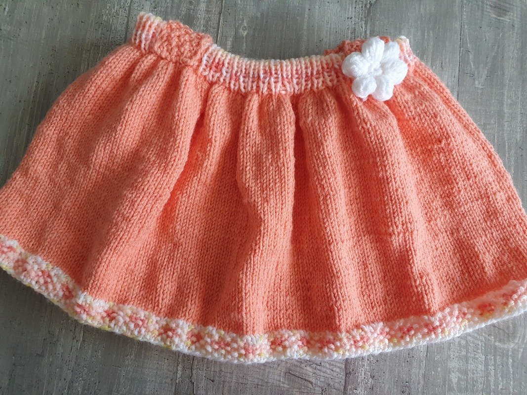 Pfirsichfarbener gestrickter Babyrock mit weißer Häkelblüte und pastellfarbener Abschlusskante