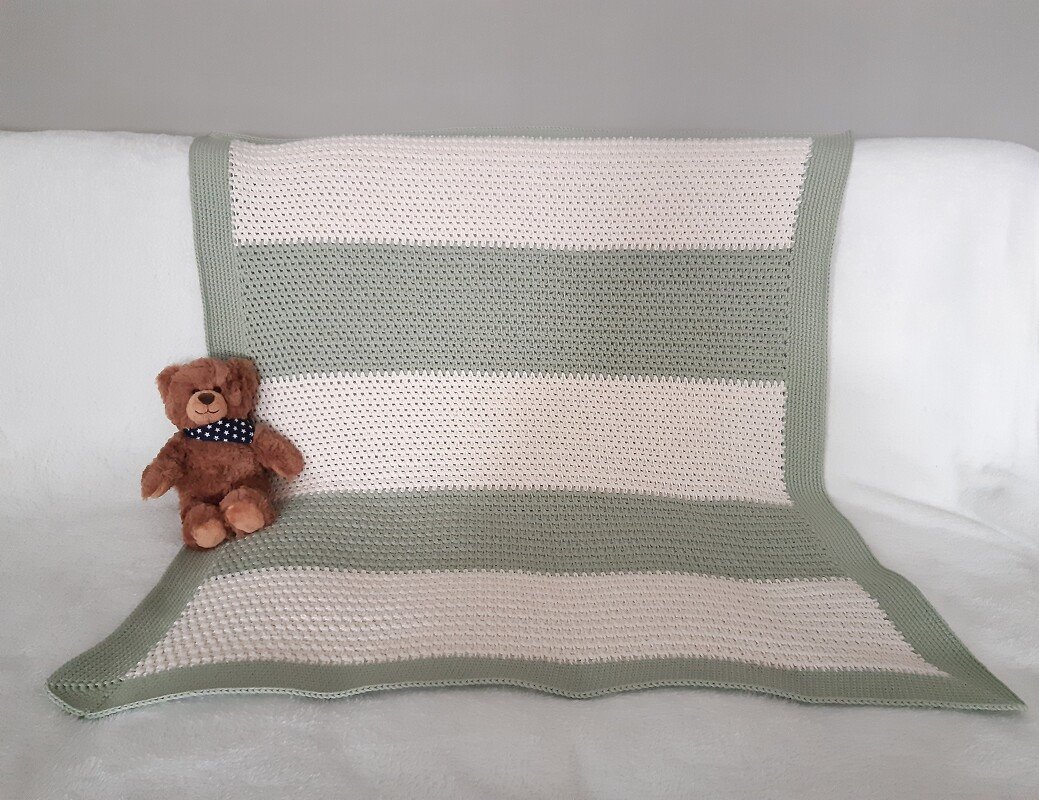 Gehäkelte Babydecke mit breiten hellgrünen und cremefarbenen Streifen, dekorativ drapiert neben einem braunen Teddybär