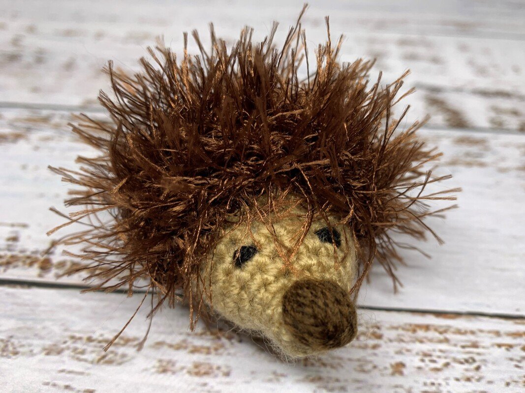 Small handcrafted crocheted hedgehog with fuzzy dark-brown yarn quills and a beige face on a whitewashed wooden surface.