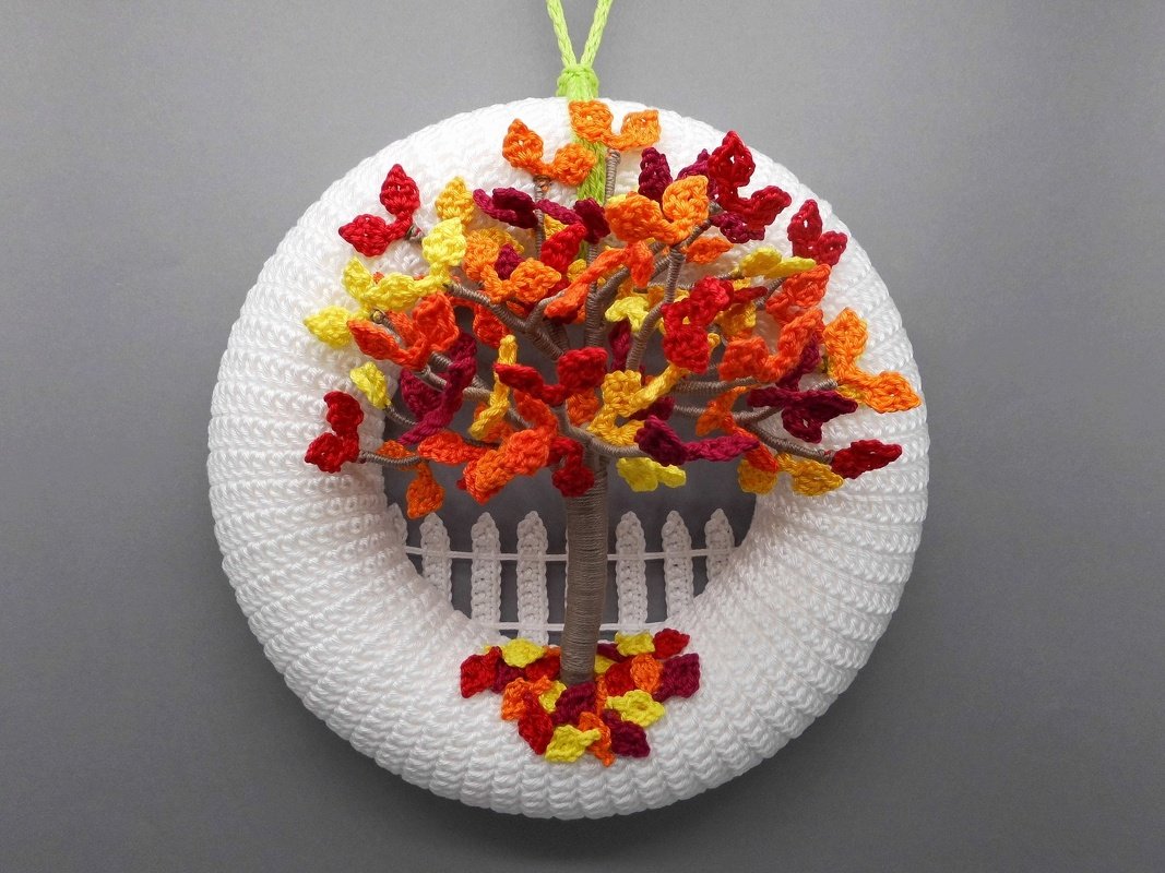 White crocheted round wreath with an autumn tree, red, orange, and yellow leaves, a small white picket fence, and fallen leaves at the base.