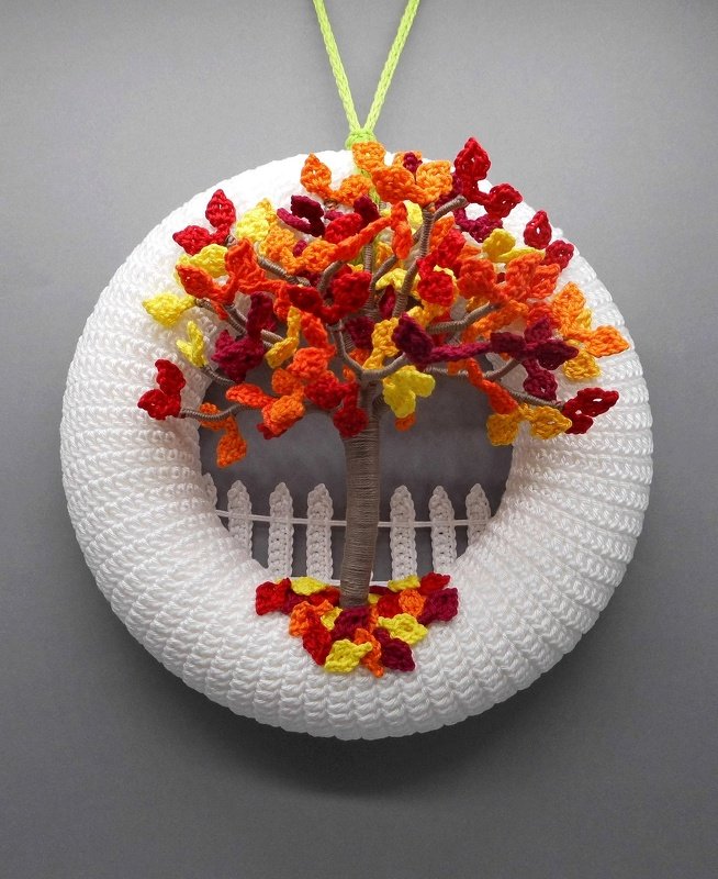 White crocheted round wreath with a central yarn-wrapped tree and red, orange and yellow crochet leaves with a small white picket fence