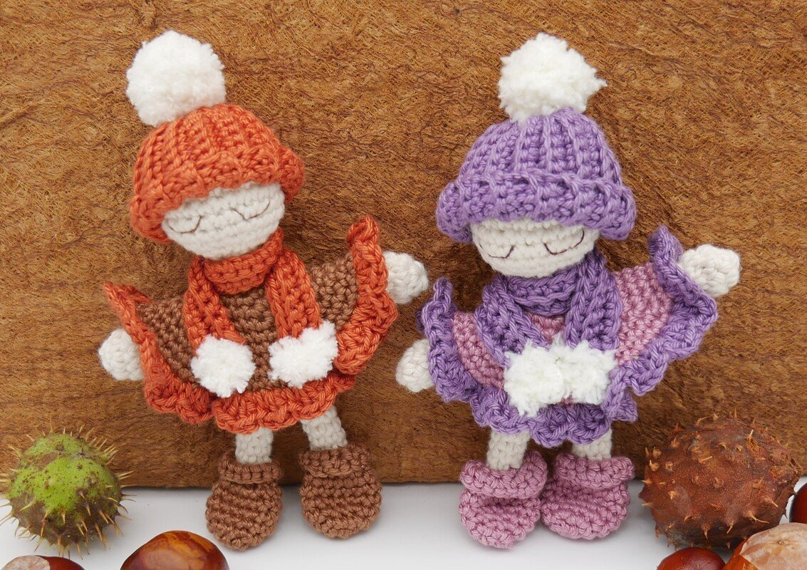 Zwei gehäkelte Amigurumi-Puppen, eine orange, eine lila, mit Mützen, Schals und Ponchos, umgeben von Kastanien vor braunem Hintergrund.