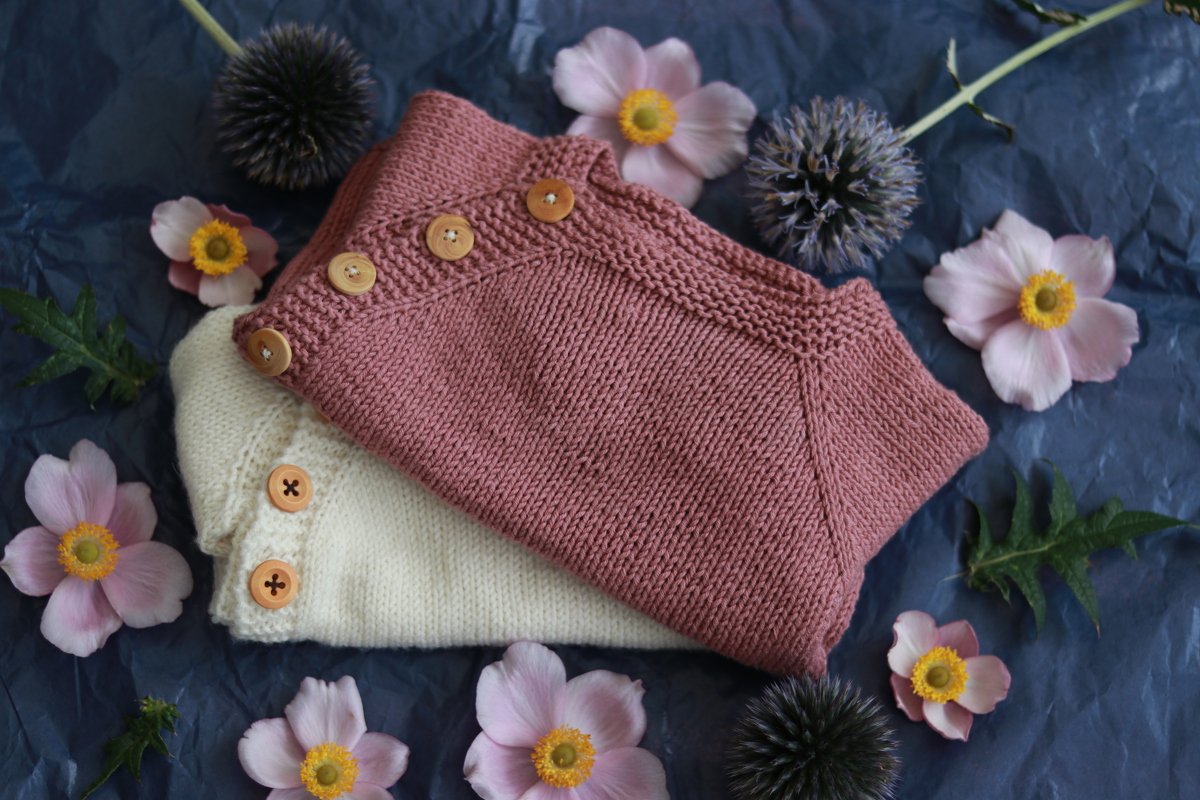 Folded dusty-rose and cream knitted baby cardigans showing raglan seam detail and wooden buttons, surrounded by flowers on dark paper.