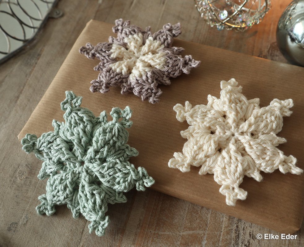 Three crocheted snowflake ornaments in cream, mint green, and two-tone beige on kraft-wrapped paper and a wooden table.