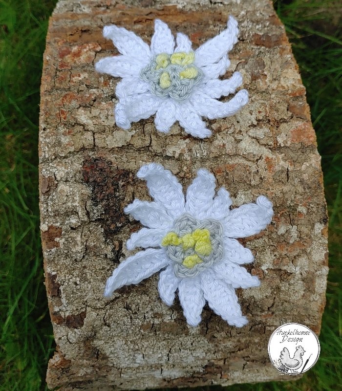 Zwei gehäkelte Edelweißblüten mit weißen Blütenblättern und hellgelb-grauen Zentren auf einem Stück Baumrinde vor Gras.