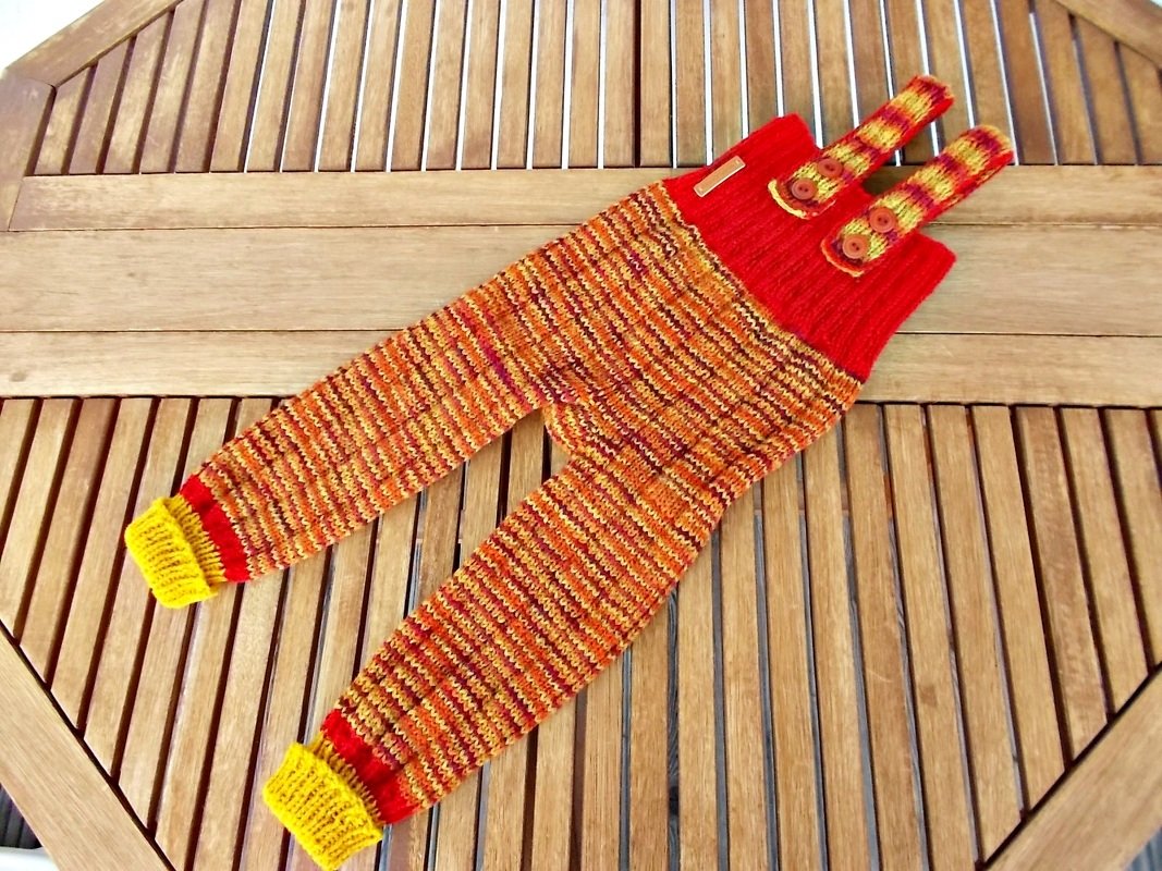 Gestrickte Kinderhose in Orange-, Rot- und Brauntönen mit gelben Bündchen und roten Knopfträgern auf Holztisch.