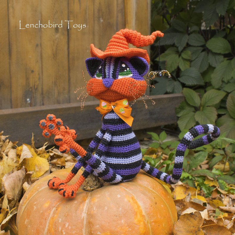 Purple and black striped crocheted cat amigurumi with an orange witch hat and bow, seated on a pumpkin.