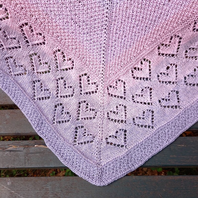 Gestricktes Dreieckstuch mit Herz-Lochmuster in Rosa- und Lilatnen auf einer Holzbank